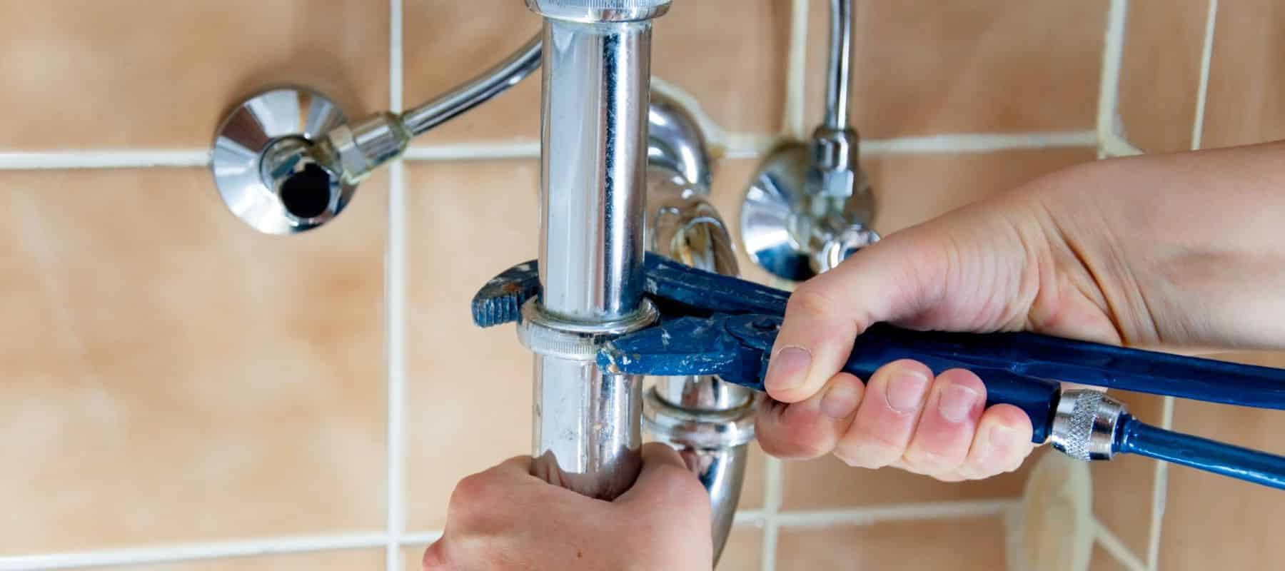 closeup of a plumber hand holding a blue wrench to tighten a section of a silver pipe under a kitchen sink