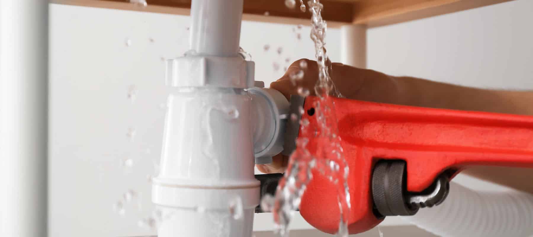 red wrench working on a white pipe under a sink that has water running out of it due to a leak