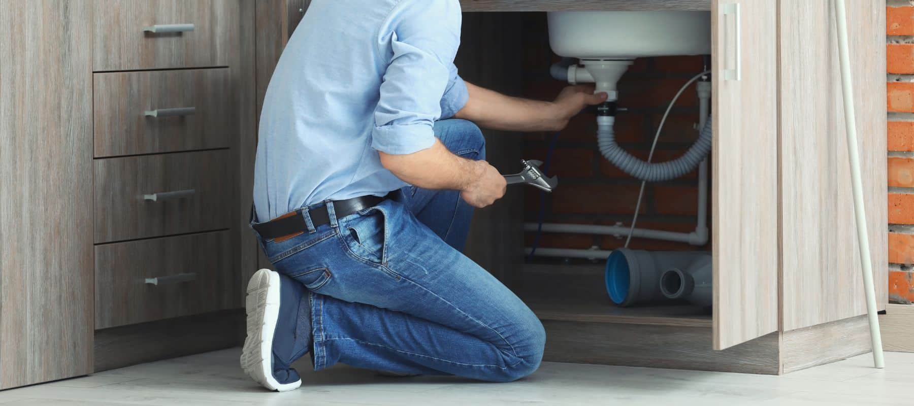 homeowner bending down in front of a bathroom sink holding a wrench to tighten a section of piping on the sink