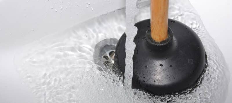 closeup of a drain in a sink with water running and a black plunger sitting next to the drain