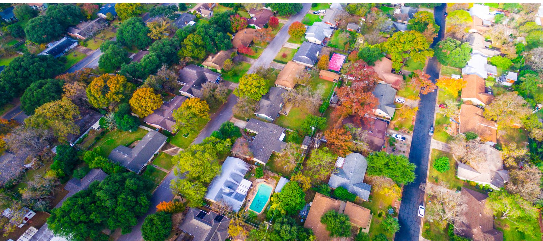 An aerial view of a suburban neighborhood showcasing an array of detached houses, each with distinct yard spaces, some featuring swimming pools. The area is interspersed with lush trees in various shades of green, orange, and yellow, suggesting a vibrant autumn season. The layout reveals a tranquil, well-spaced residential area, with roads gently curving through the landscape
