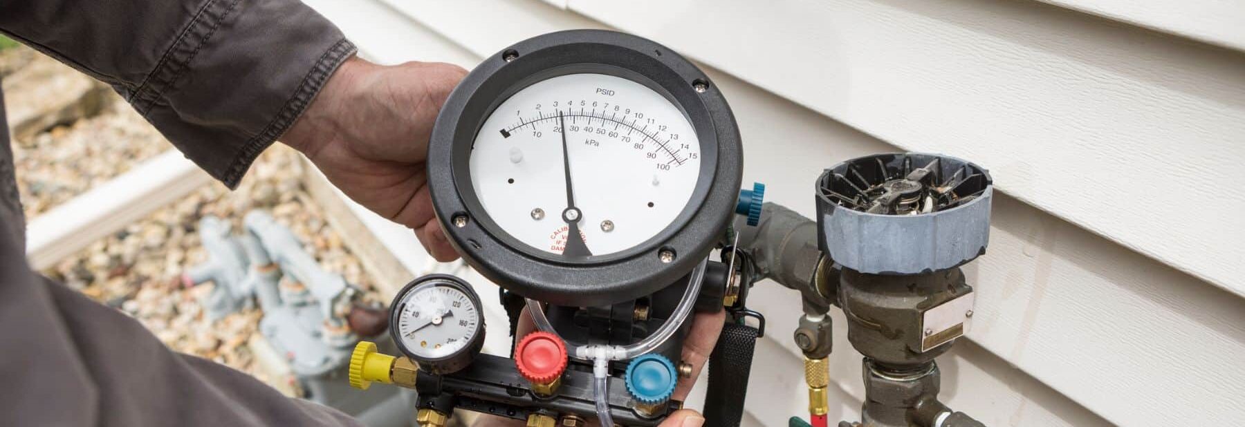 A technician's hand holding a pressure gauge device near an outdoor gas meter setup on the side of a house. The gauge shows the pressure reading and is connected to various hoses and fittings, indicating a gas line inspection or maintenance work. The background includes gravel and part of the house's exterior wall, suggesting an outdoor work environment