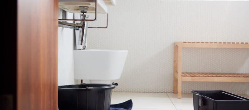 Under-sink area in a bathroom with plumbing exposed, a black bucket placed underneath, and a wooden bench nearby, indicating a potential leak or plumbing maintenance