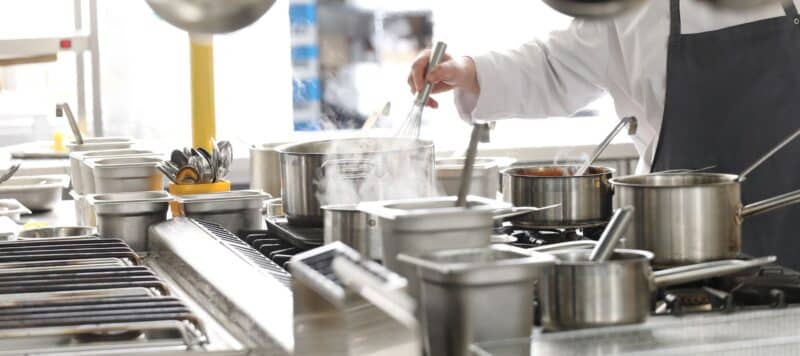 A chef stirs a steaming pot on a busy commercial kitchen stove with various pans and utensils in the background