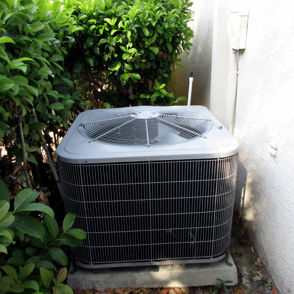 An outdoor air conditioning unit placed near green shrubs, partially concealed by surrounding vegetation