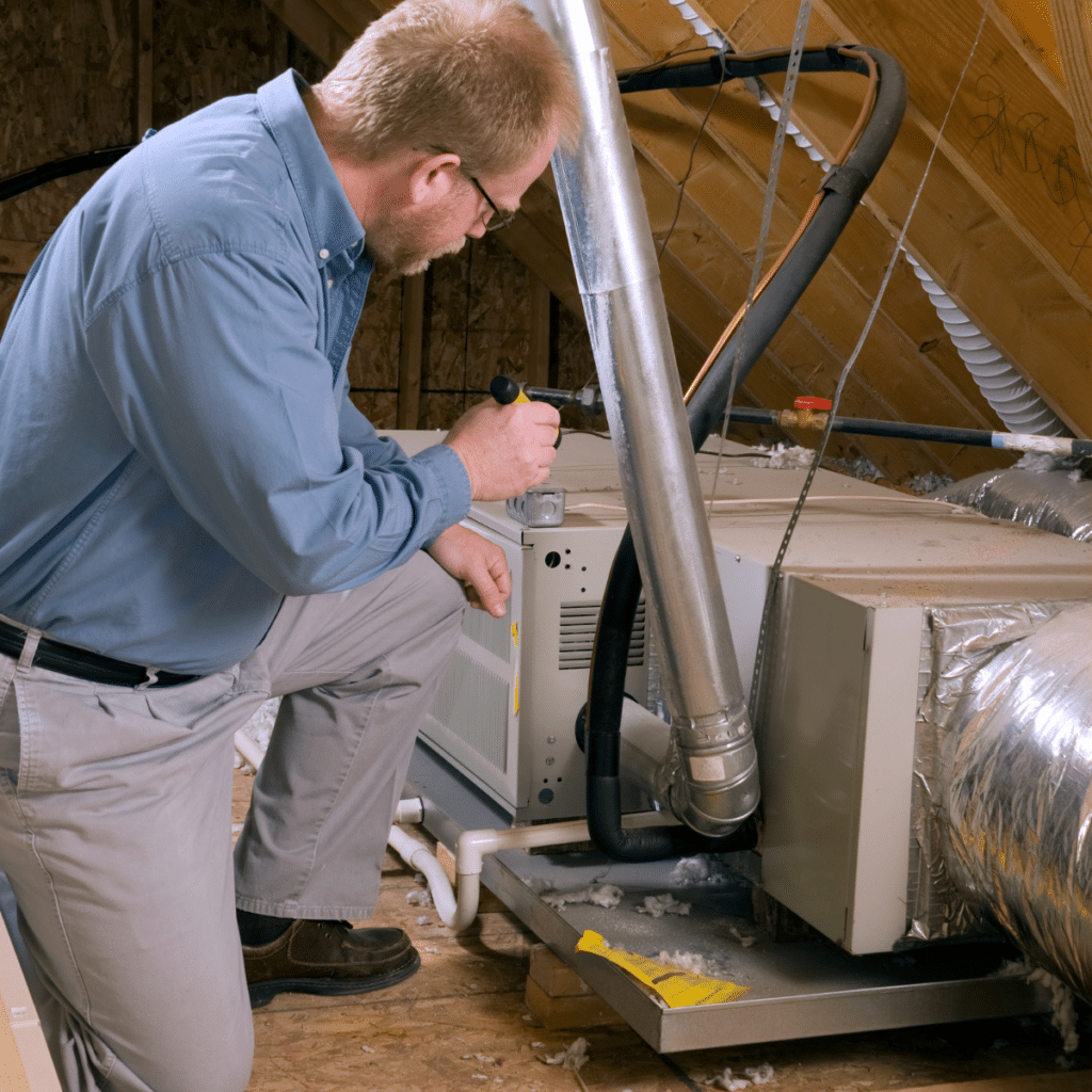 Technician kneeling next to indoor HVAC equipment in an attic, conducting a system inspection