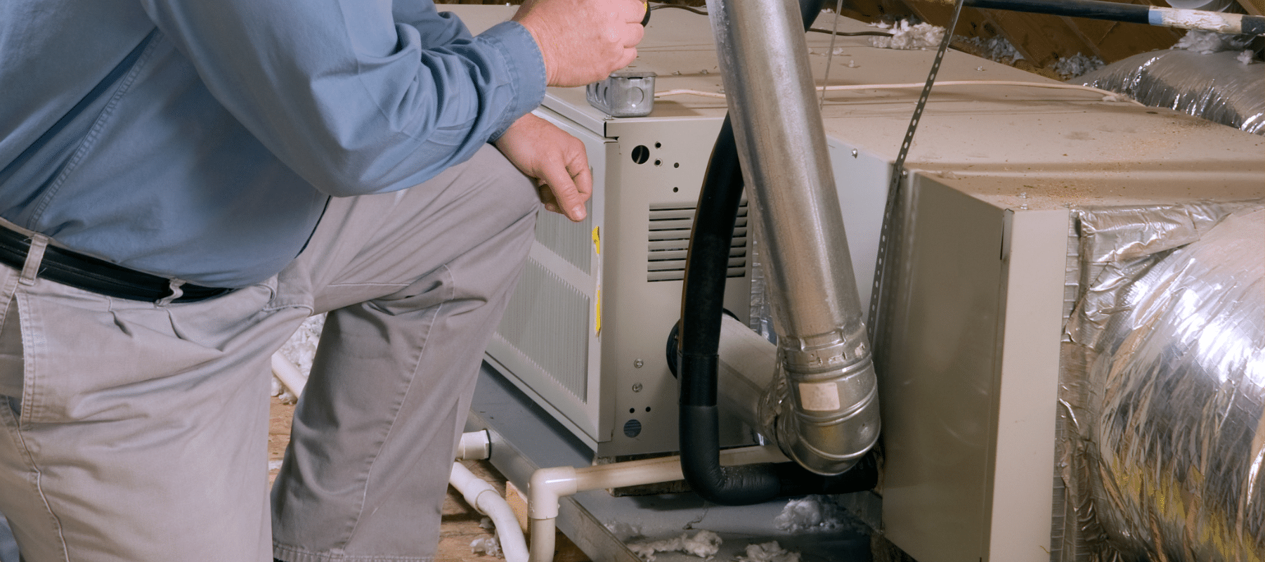 Technician kneeling next to indoor HVAC equipment in an attic, conducting a system inspection