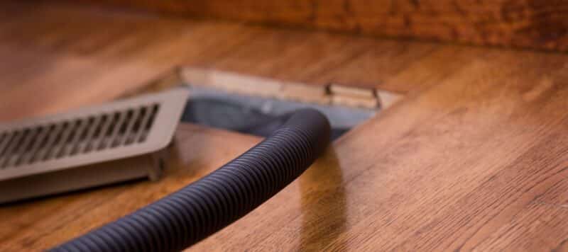 Close-up of a vacuum hose cleaning out a floor air vent on a wooden surface, with the vent cover removed