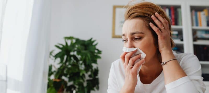 Woman holding a tissue to her nose, looking unwell while sitting indoors.